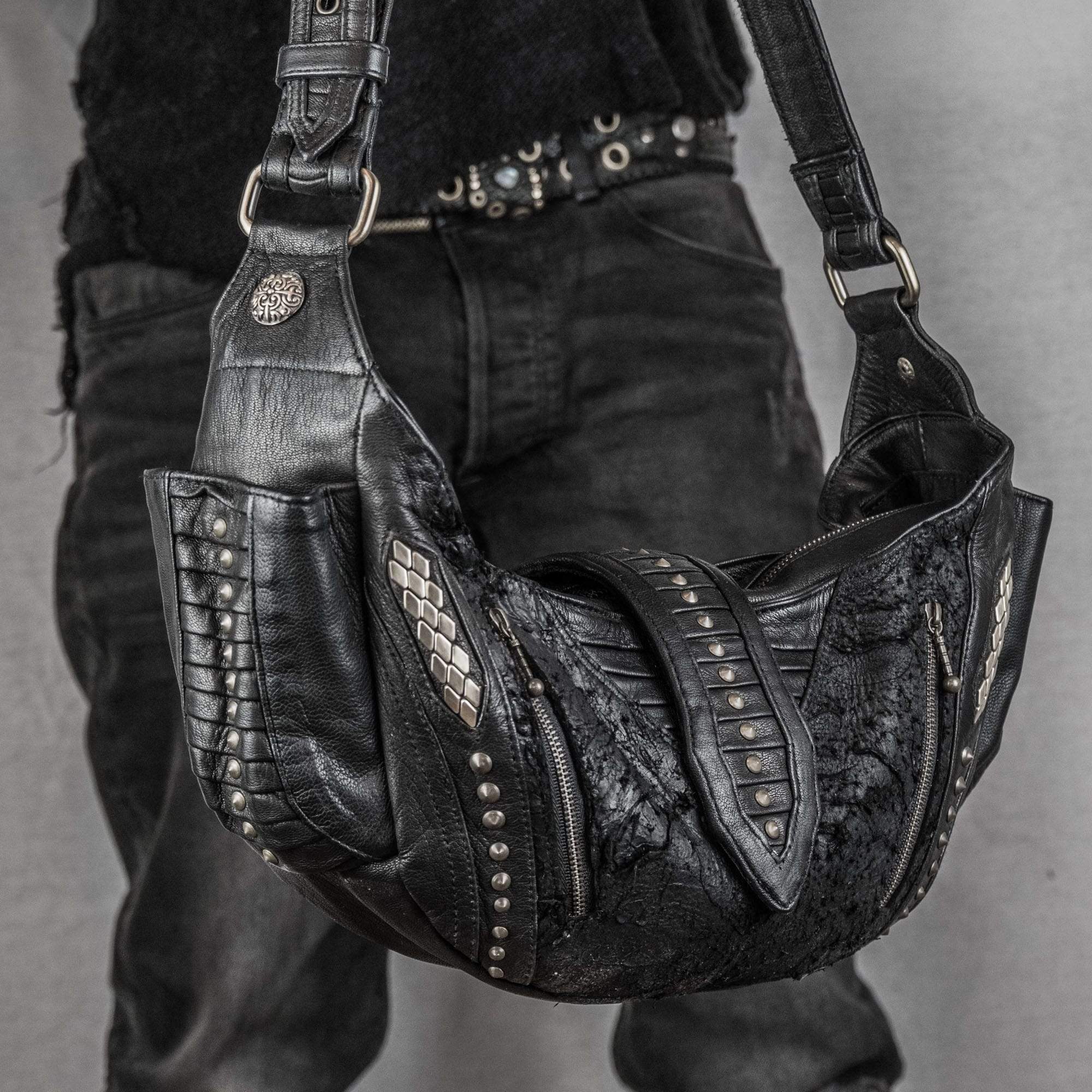 A person dressed in dark clothing holds a Nebula Unisex Leather Off Shoulder Cross Body HandBag by Kaya Movement. This custom black leather bag boasts intricate details, including metallic studs, textured patterns, and side panels akin to reptile skin. The individual's belt also features metal studs, perfectly complementing the edgy design of the handbag, making it ideal for dystopian festivals.